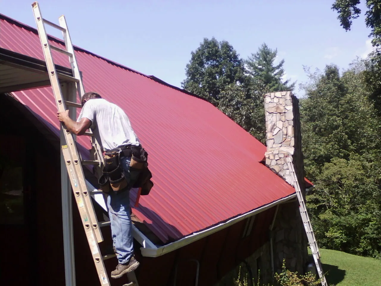Roofer on a ladder beside a red metal roof and stone chimney — Soffit & Fascia Repair by Roofing Brothers in Lahoma